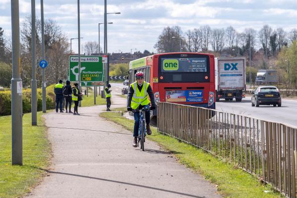 Wide shared cycle path next to busy road. Man cycling towards the camera on path. People in background are waiting for a red bus which is approaching them. Large green Kettering route sign near them. Trees and blue sky in background,